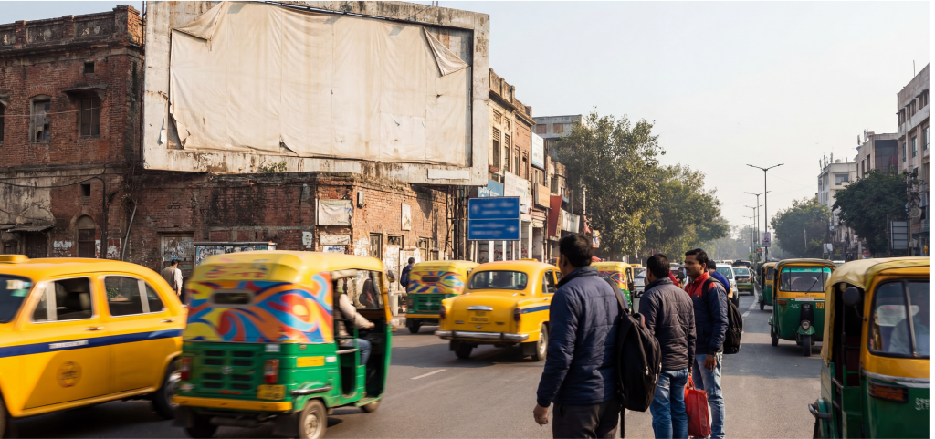 Busy Indian street with yellow taxis, auto-rickshaws, pedestrians, and an old building featuring a blank billboard.